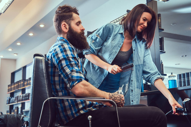 Handsome stylish bearded male with a tattoo on arm dressed in a flannel shirt holding juice while getting a haircut. - Foto, afbeelding