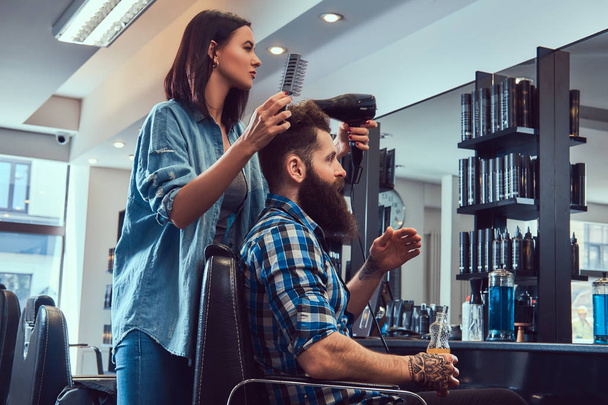 Гарний стильний бородатий чоловік з татуюванням на руці, одягнений у фланелеву сорочку, що тримає сік, тоді як перукарня-жінка використовує фен у перукарні
. - Фото, зображення