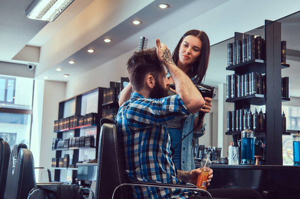 Красивый стильный бородатый мужчина с татуировкой на руке, одетый во фланелевую рубашку, держа сок, в то время как парикмахерша использует фен в парикмахерской
. - Фото, изображение