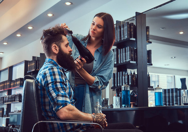 Гарний стильний бородатий чоловік з татуюванням на руці, одягнений у фланелеву сорочку, що тримає сік, тоді як перукарня-жінка використовує фен у перукарні
. - Фото, зображення