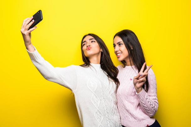 Portrait of two cheerful girls dressed in sweaters standing and taking a selfie isolated over yellow background - Φωτογραφία, εικόνα