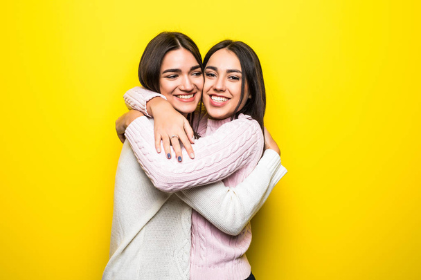 Portret van twee gelukkige meisjes gekleed in truien knuffelen geïsoleerd op gele achtergrond - Foto, afbeelding