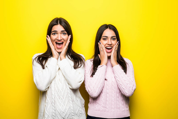 Két izgatott lány öltözött pulóverek, sikoltozik és látszó-on fényképezőgép, izolált át a sárga háttér portréja - Fotó, kép