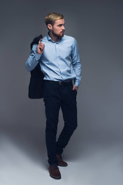 Stylish and professional. Fashionable entrepreneur holding his jacket on the shoulder and put the other hand into the pocket in front of grey background. - Photo, Image