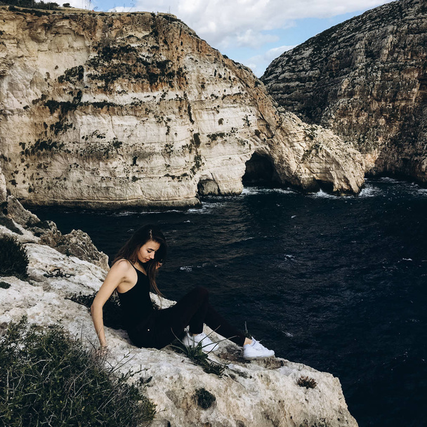 vista de mujer joven con pelo largo en ropa negra en las montañas rocosas en la costa con agua oscura en el día ventoso
 - Foto, Imagen