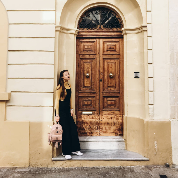 Woman wearing black romper, yellow jumper and white sneakers standing near house entrance and holding pink backpack in hand. - Фото, изображение