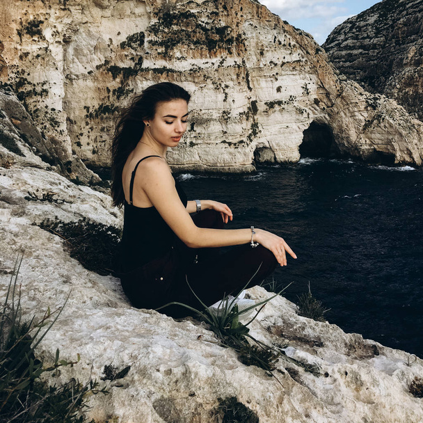 Blick auf junge Frau mit langen Haaren und geschlossenen Augen in schwarzer Kleidung auf felsigen Bergen an der Küste mit dunklem Wasser bei windigem Tag - Foto, Bild