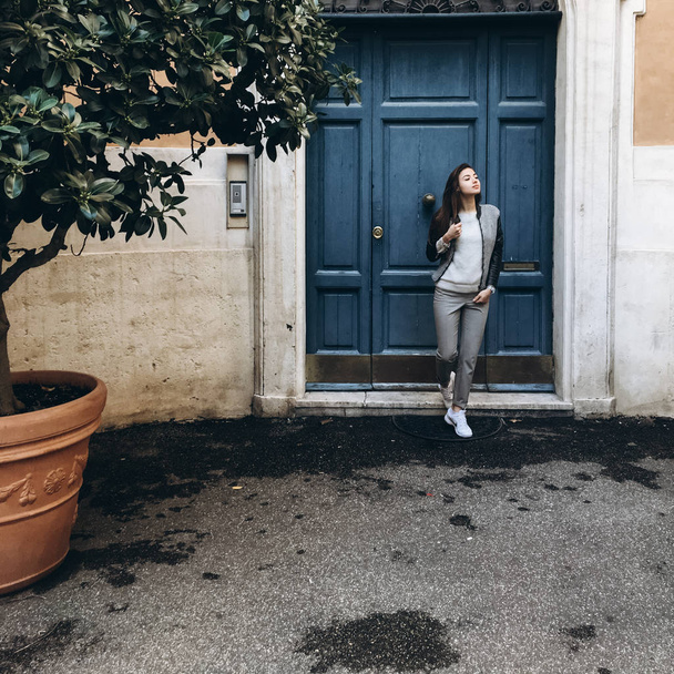 näkymä nuori nainen pitkät hiukset aiheuttaa lähellä sininen ovi antiikin rakennuksen vihreä puu
 - Valokuva, kuva