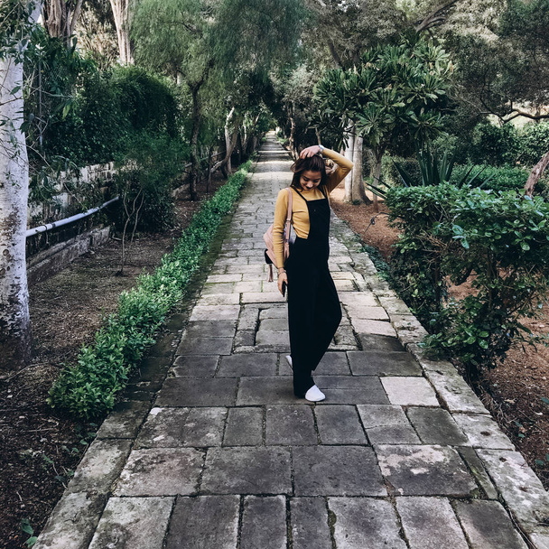 Woman wearing black romper, yellow jumper and white sneakers walking on alley and playing with hair in old garden. - Photo, Image