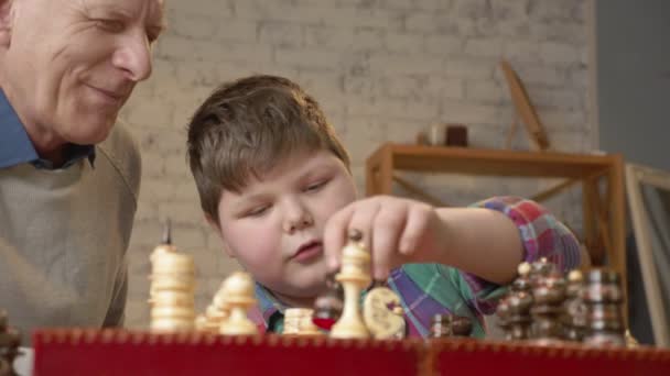 Grandfather and grandson plays chess. Elderly man teaches chess to play a young fat child. Home comfort, family idyll, cosiness concept, difference of generations, close up. 60 fps - Felvétel, videó