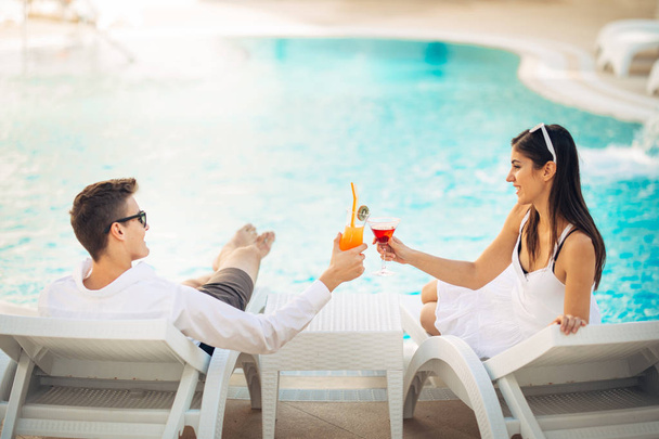 Positive happy couple having a romantic afternoon by the pool in luxury summer vacation resort.Drinking cocktails.Relaxing and enjoying spa wellness weekend.Travel, honeymoon,relationship concept - Foto, afbeelding