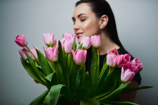 Young beautiful happy girl holding spring bouquet of tulips, posing on white background.  Portrait of attractive young woman with tulips is standing in light room and smiling. Happy international women's day! Celebrating 8th of March. - Foto, Imagen