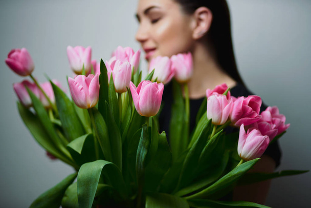 Young beautiful happy girl holding spring bouquet of tulips, posing on white background.  Portrait of attractive young woman with tulips is standing in light room and smiling. Happy international women's day! Celebrating 8th of March. - Фото, изображение