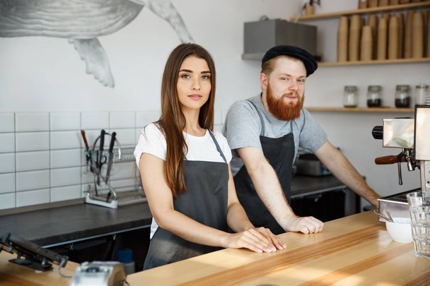Koffie businessconcept - positieve bebaarde man en mooi aantrekkelijke jongedame barista paar in schort camera terwijl staande aan de toog te kijken - Foto, afbeelding