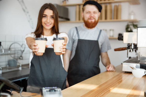 Coffee Business Concept - Positive young bearded man and beautiful attractive lady barista couple giving take away cup of coffee to custome at the modern coffee shop - Photo, Image