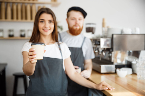 Coffee Business Concept - Positive young bearded man and beautiful attractive lady barista couple giving take away cup of coffee to custome at the modern coffee shop - Fotó, kép