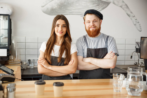 Coffee Business Concept - Positiivinen nuori parrakas mies ja kaunis viehättävä nainen barista pari esiliina katselee kameraa seistessään baaritiskillä
. - Valokuva, kuva