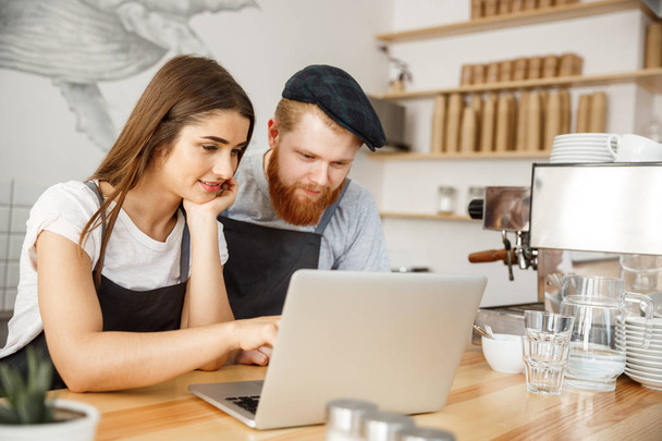Coffee Business Concept - Los propietarios satisfechos y sonrientes buscan en la computadora portátil para pedidos en línea en la cafetería
 - Foto, imagen