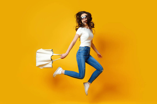 Beautiful young woman in sunglasses, white shirt, blue jeans jumping with bag on the yellow background - Foto, afbeelding