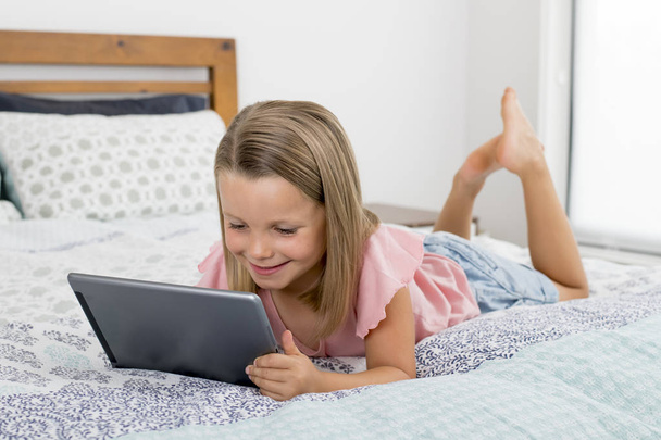 甘いと美しいブロンド 6 または 7 歳少女はインターネットを用いたデジタル タブレット パッド見ていて楽しい幸せな笑みを浮かべてベッドに横になっています。 - 写真・画像