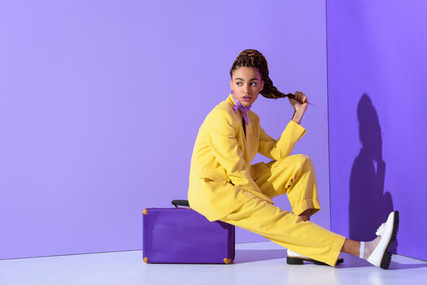 Afro-Amerikan kız mor bavul, trendy ultra violet arka plan üzerinde oturan sarı elbiseli poz - Fotoğraf, Görsel