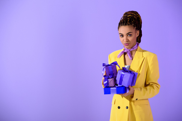 красивая африканская американка в желтом костюме держит подарочные коробки, на модном ультрафиолетовом фоне
 - Фото, изображение
