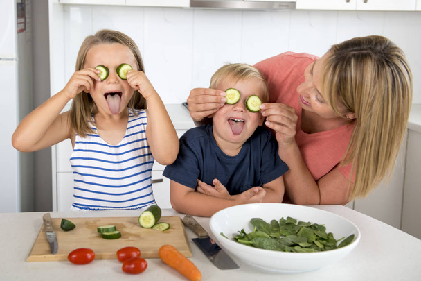 молода приваблива жінка готує разом з маленьким сином 3 і 7 років і дочкою, граючи щасливою з огірковим стрижнем, як очі готують салат
 - Фото, зображення