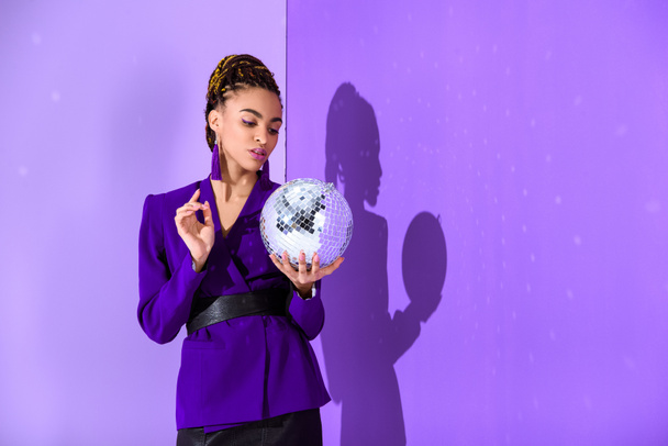 elegantes afrikanisch-amerikanisches Mädchen in lila Jacke mit Discokugel an der ultravioletten Wand  - Foto, Bild