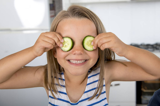 young beautiful and adorable girl 6 or 7 years old having fun at home kitchen putting cucumber slices on her eyes smiling happy - Photo, image