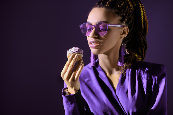 Stylish african american girl in ultra violet jacket and sunglasses with cupcake, isolated on dark purple - Photo, Image