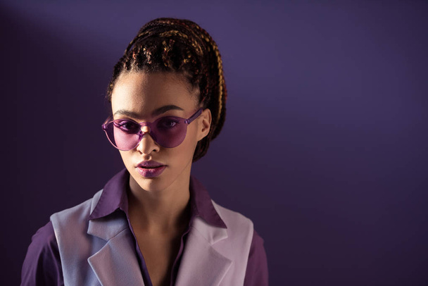 Fashionable african american girl posing in purple sunglasses, isolated on purple - Foto, Imagem