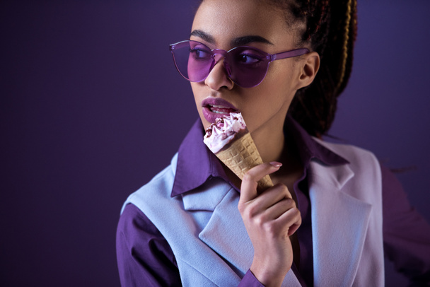 Chica afroamericana posando en gafas de sol púrpura con helado en cono, aislado en púrpura
 - Foto, Imagen