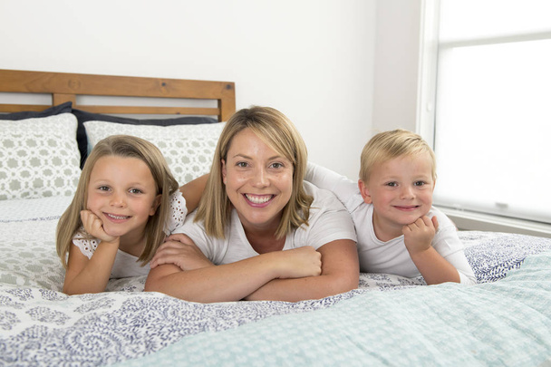 jonge blonde Kaukasische vrouw liggend op bed samen met haar kleine zoete 3 en 7 jaar oude zoon en dochter glimlachend speels en blij - Foto, afbeelding