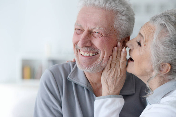 Close-up portrait of senior woman whispering in her husbands ear at home - 写真・画像