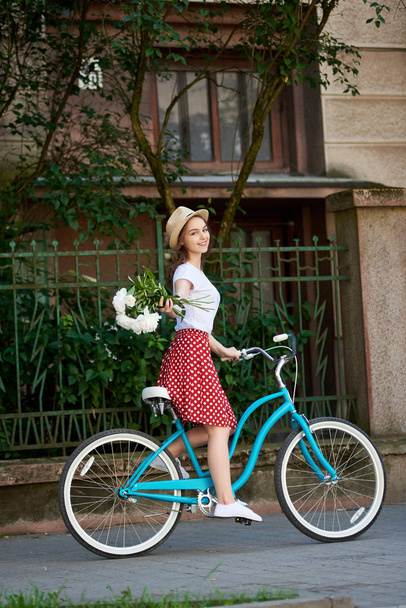 Tender young woman in a red skirt with polka dots and a hat on her head on a blue retro bike with peonies.  - Foto, Imagen