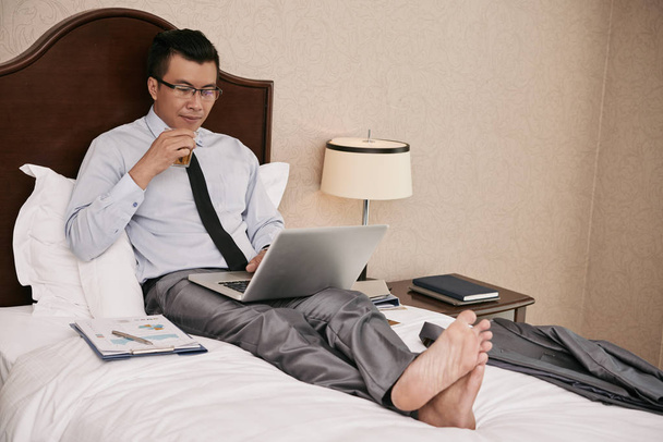 Empresario vietnamita disfrutando de un vaso de whisky cuando trabaja en el portátil en la cama
 - Foto, imagen