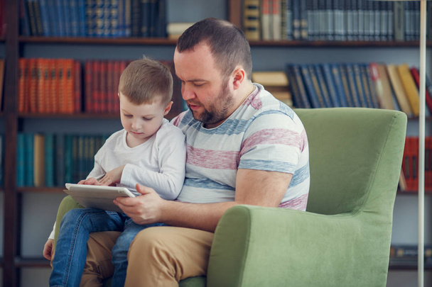 Image of happy father and young son sitting with tablet on green chair - Foto, Bild