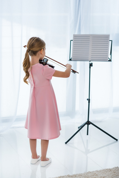Rückansicht eines kleinen Kindes im rosa Kleid, das zu Hause Geige spielt - Foto, Bild