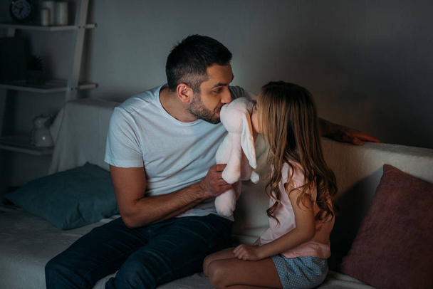 Vater und kleine Tochter küssen Spielzeug, während sie zu Hause auf dem Sofa sitzen - Foto, Bild