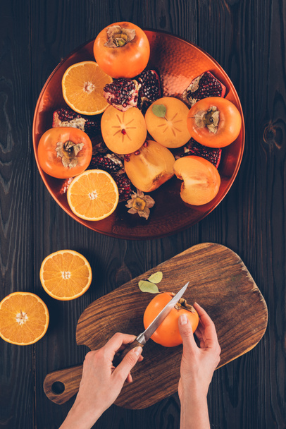 cropped image of woman cutting persimmon on wooden board - Φωτογραφία, εικόνα