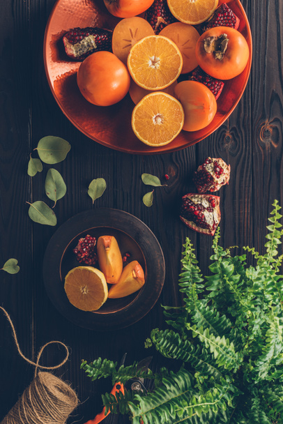top view of persimmons with cut oranges and pomegranates on plates on wooden table - Photo, Image