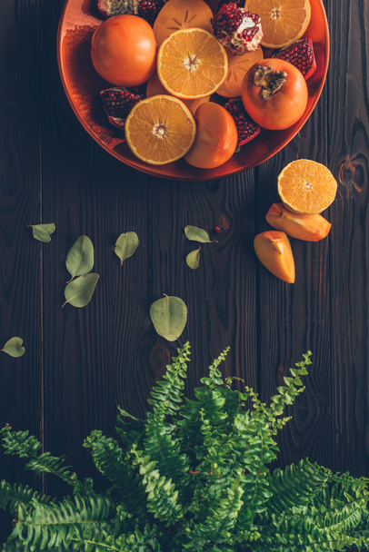 カット オレンジ、ザクロ、柿のトップ ビュー プレートとテーブルの上の緑の植物 - 写真・画像