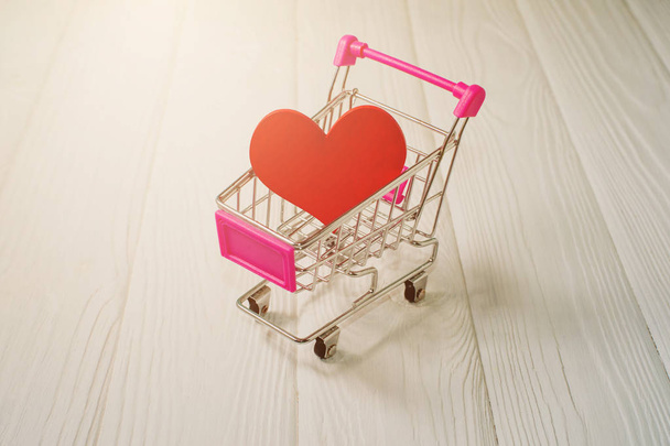 Red heart in a mini supermarket trolley on a white wooden table. Love shopping concept. Discounts and sales on a black Friday. Gift on Valentine's Day. - 写真・画像