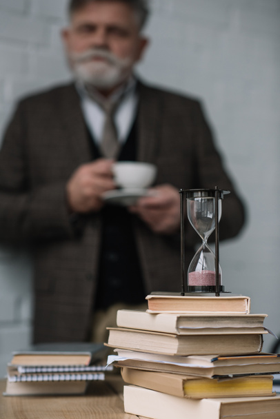 hämärtynyt vanhempi mies juo kahvia pino kirjoja ja tiimalasi etualalla
 - Valokuva, kuva