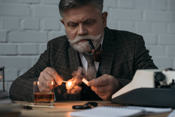 vousatý senior spisovatel s hořící zápas kouření potrubí na pracovišti - Fotografie, Obrázek