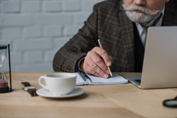 Schnappschuss eines älteren Schriftstellers, der mit Laptop arbeitet und Notizen in Notizbuch macht - Foto, Bild