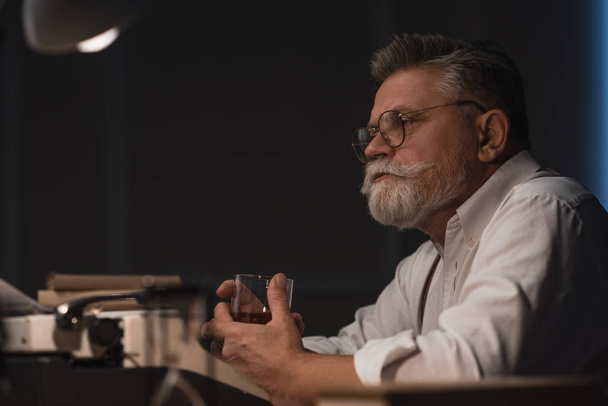 вдумливий старший письменник сидить на робочому місці зі склянкою віскі ізольовані на чорному
 - Фото, зображення
