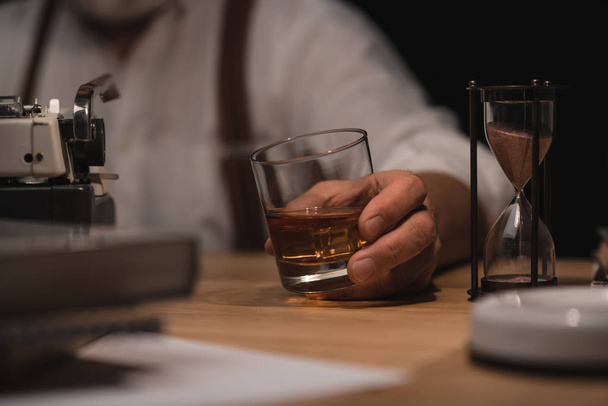 recortado tiro de escritor senior con vaso de whisky en la mano en el lugar de trabajo
 - Foto, Imagen