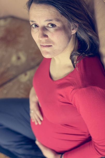 werdende Mutter in rotem Hemd lässig auf einer Couch sitzend. - Foto, Bild
