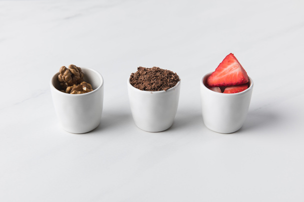 Крупный план трех чаш с грецкими орехами, тертым шоколадом и клубникой на сером
 - Фото, изображение